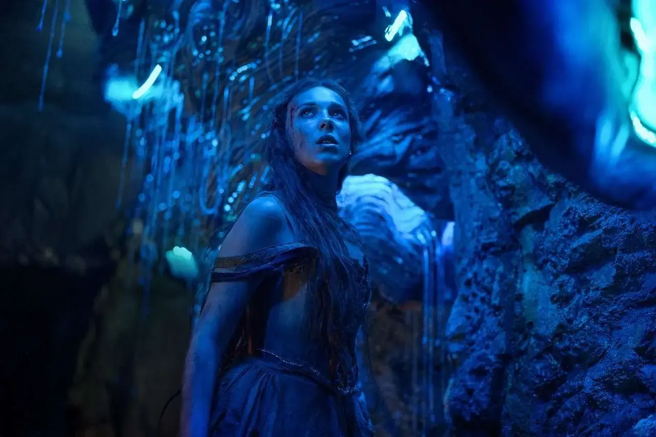 Una giovane donna con lunghi capelli scuri, vestita con abiti blu, guarda verso l'alto in una grotta illuminata da luci blu. La sua espressione è di stupore e meraviglia, quasi come se fosse una damigella in pericolo in un mondo alieno. La recensione d...