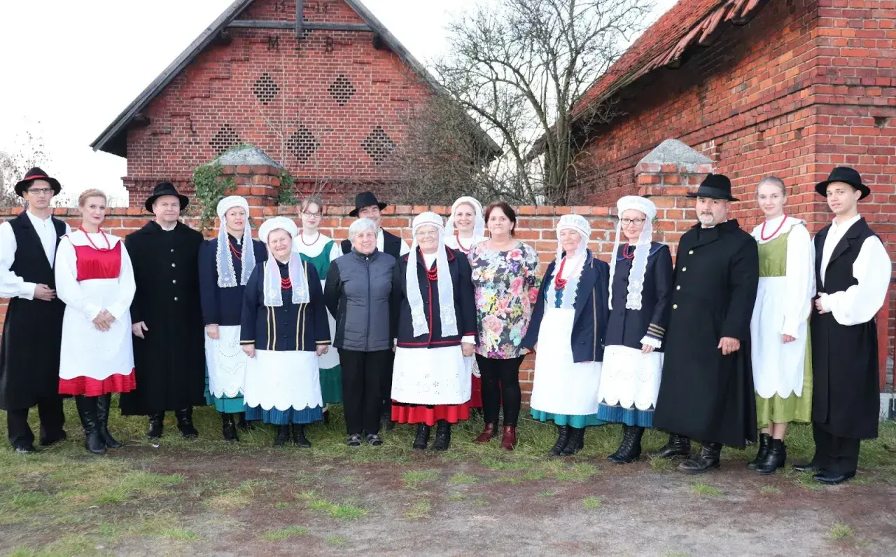 Grupa osób w tradycyjnych strojach wielkopolskich pozuje przed ceglanym budynkiem.