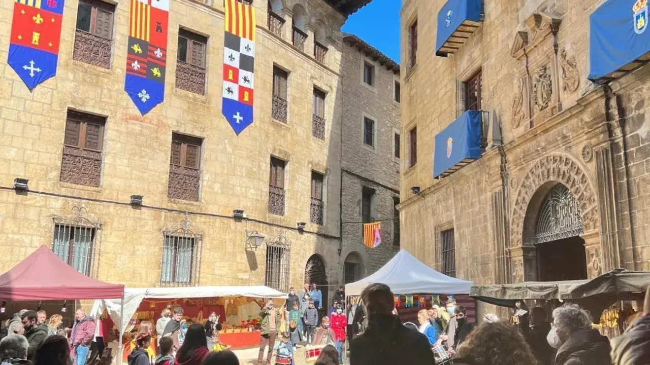 Gente disfruta del mercado medieval de las tres culturas, con puestos y banderas históricas adornando edificios antiguos.