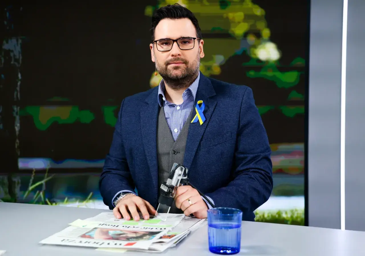 Michał Szajder, dziennikarz, w garniturze z przypinką w barwach Ukrainy, siedzi przy stole z gazetami i szklanką wody.