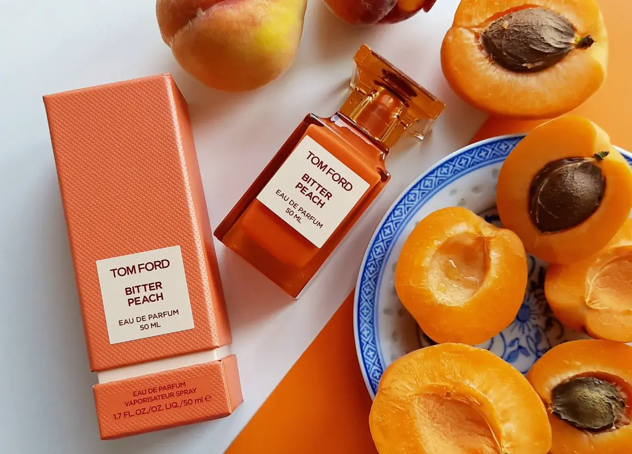 Pomarańczowe opakowanie i buteleczka perfum Tom Ford Bitter Peach otoczone soczystymi brzoskwiniami.