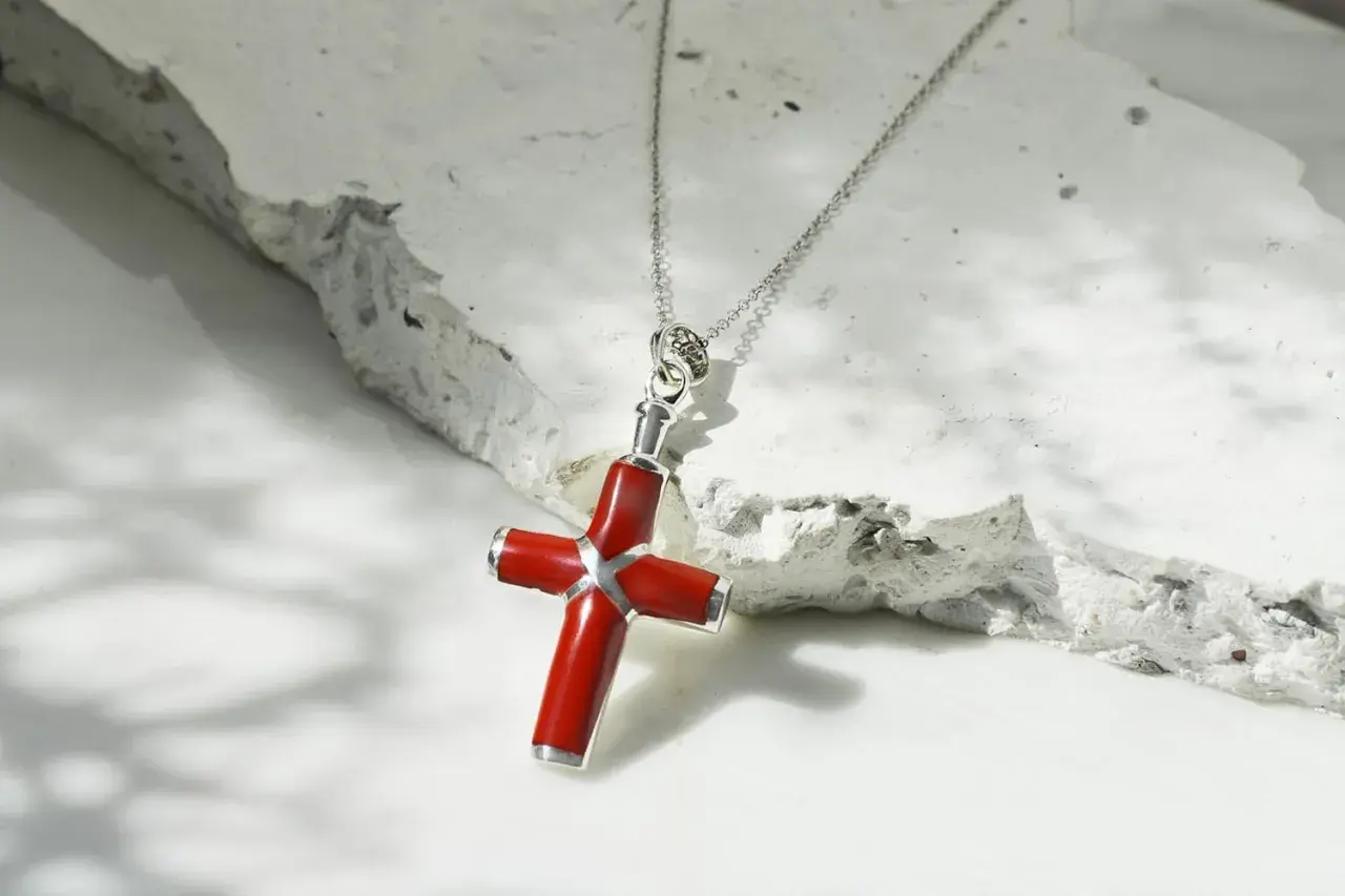 Collar con colgante de cruz de coral rojo y plata, sobre una superficie texturizada de piedra blanca.