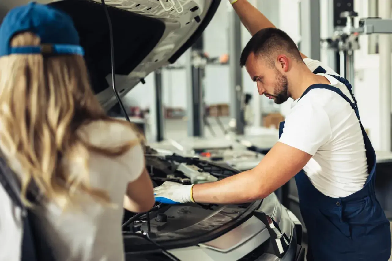 Mechanik z rękawiczkami bada silnik, szukając przyczyny usterki systemu zarządzania silnikiem. Obok stoi kobieta w czapce.