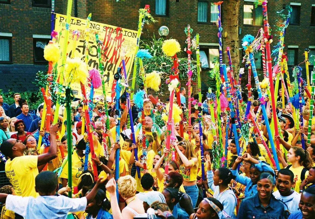 Gente celebra en un desfile, levantando coloridos bastones adornados con pompones y cintas. Un cartel dice "POLE-O-NG STATION". Parece una fiesta, quizás un bank holiday.