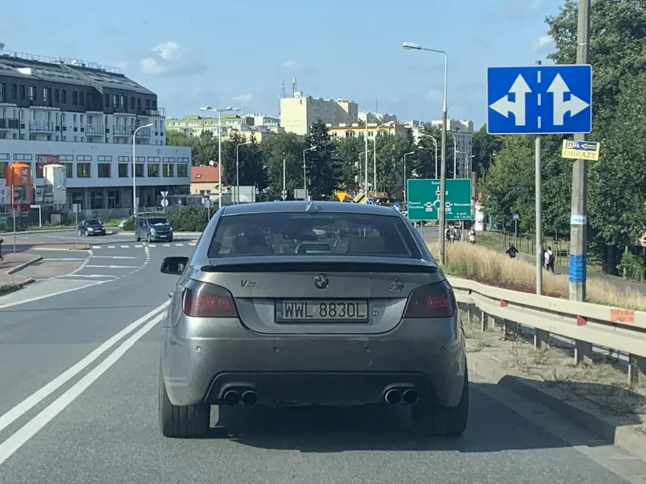 Szare BMW z rejestracją WWL 8830L na drodze, w tle budynki i znaki drogowe.