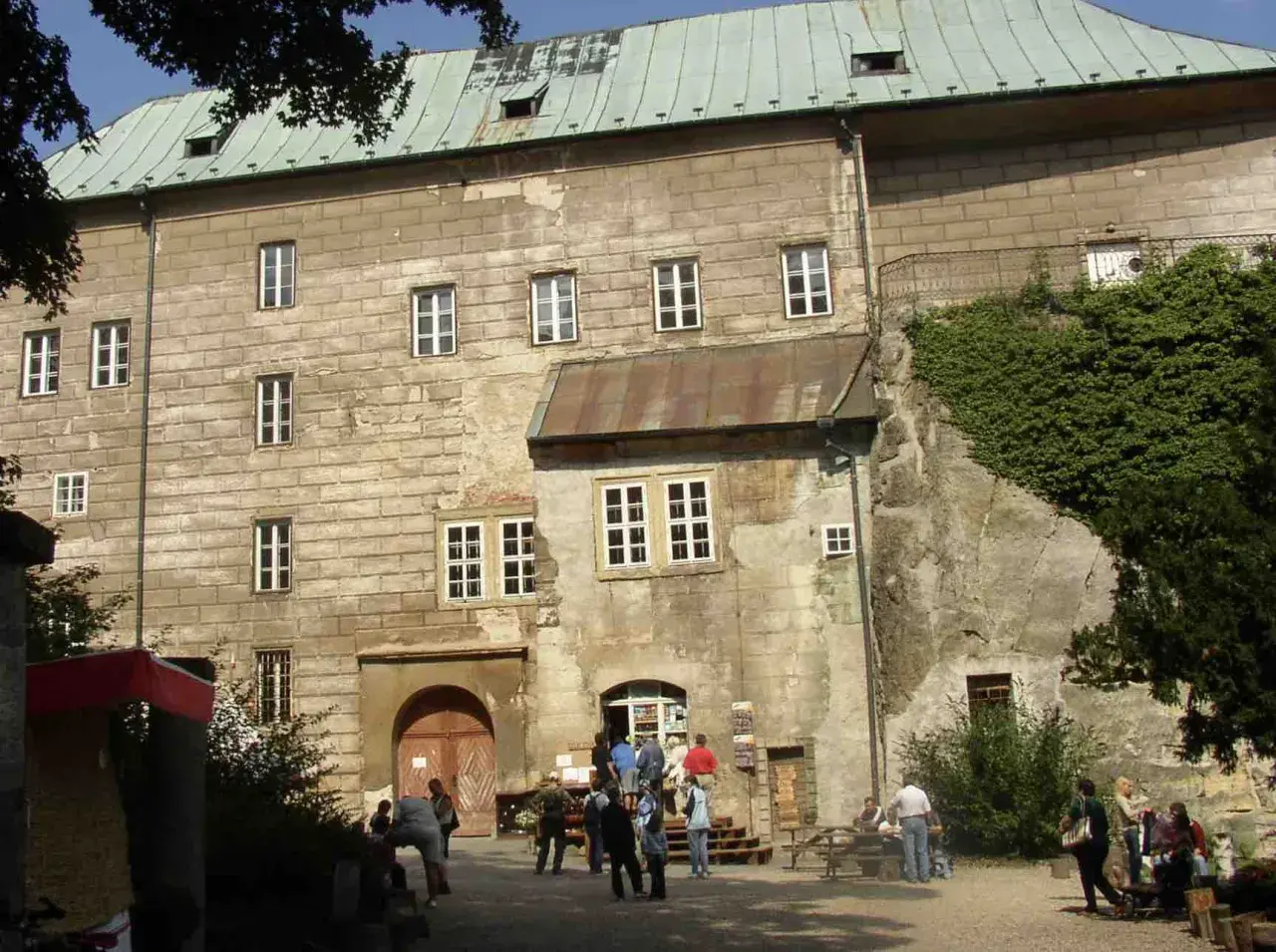 Kamienny zamek Houska z zielonym dachem i licznymi oknami, otoczony zielenią.