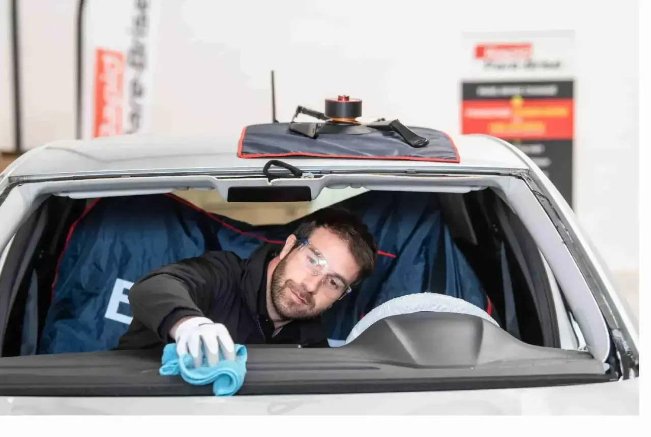 Un technicien porte des lunettes de protection et utilise un chiffon bleu pour nettoyer l'intérieur du pare-brise d'une voiture.