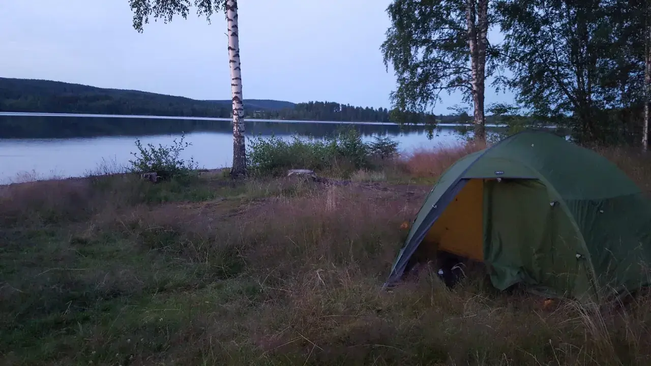 En grön campingtält står vid en sjö, med en skogklädd kulle i bakgrunden. En perfekt plats för äventyr längs 7 torpsleden.