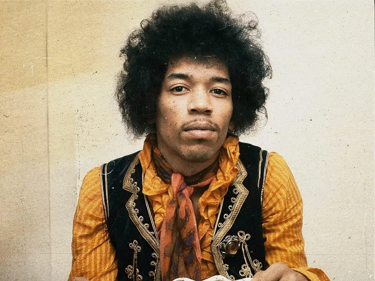 Jimi Hendrix w pomarańczowej koszuli i kamizelce, z charakterystyczną fryzurą. W tle subtelna, lekko zatarte tło, jakby w **purple haze**.