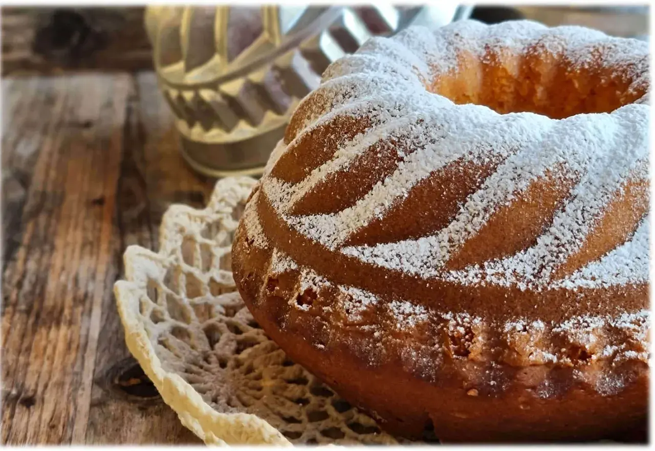 Ciambellone soffice come una nuvola, cosparso di zucchero a velo, adagiato su un centrino di pizzo, su un tavolo di legno rustico.