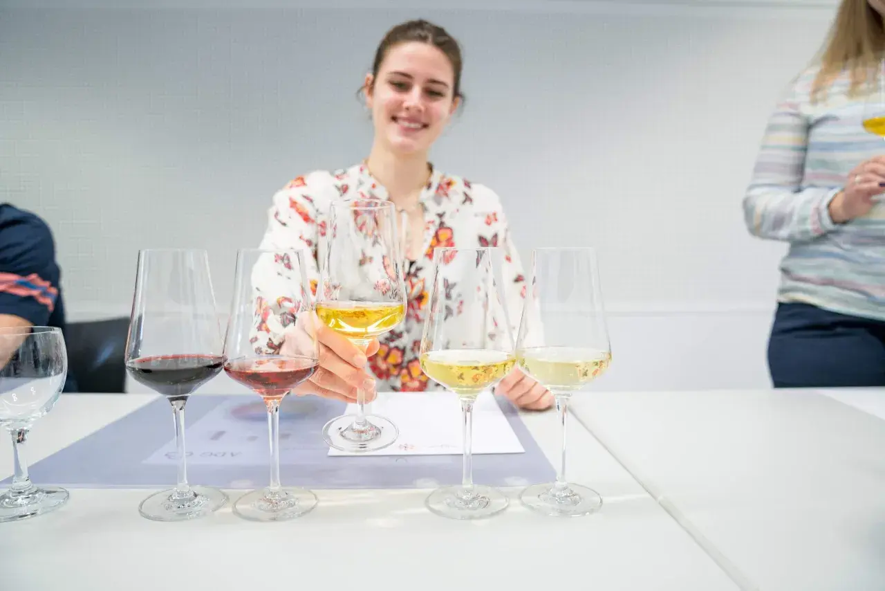 Eine Frau mit Blumenmuster-Bluse verkostet Wein, ein Schritt auf dem Weg, Sommelier zu werden. Mehrere Gläser mit Rot-, Rosé- und Weißwein stehen auf dem Tisch.