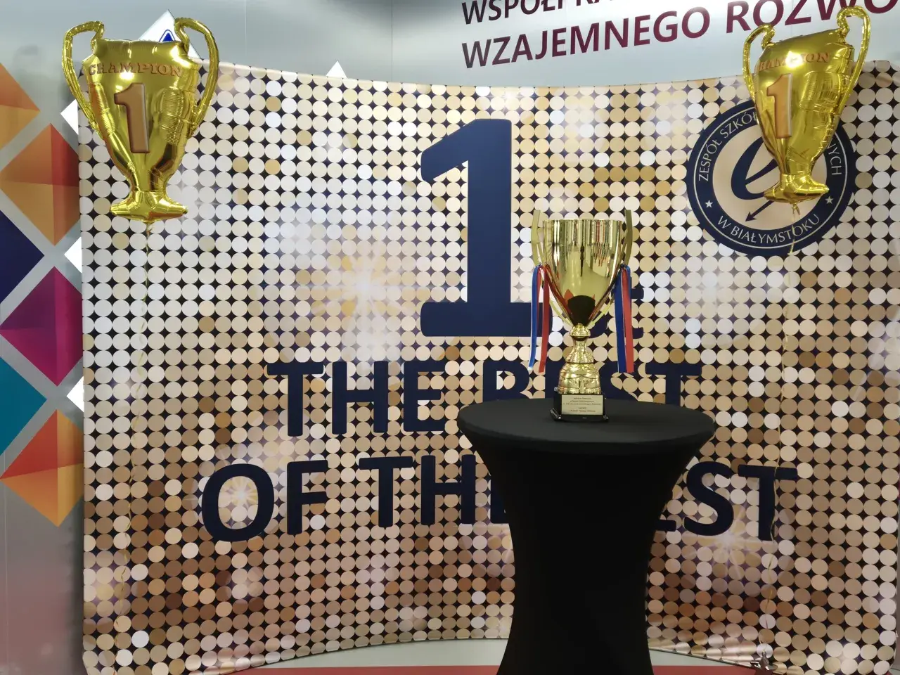 Złote puchary i balon w kształcie pucharu na tle błyszczącej ścianki z napisem "THE BEST OF THE BEST".