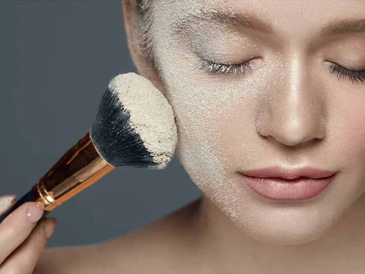 Applicazione di cipria per il baking trucco su un viso femminile. Un pennello grande raccoglie la polvere per un finish impeccabile.