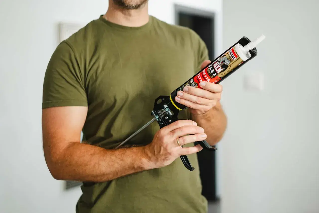 Mężczyzna w zielonej koszulce trzyma pistolet z tubą kleju do listew przypodłogowych plastikowych.
