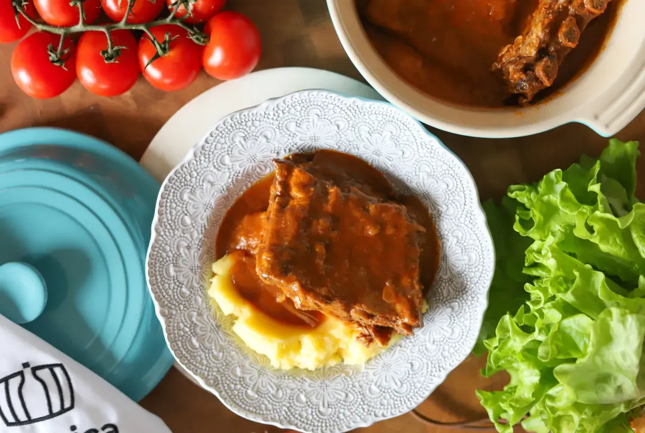 Pyszny sos z żeberek podany z purée ziemniaczanym na ozdobnym talerzu. Obok świeża sałata i pomidory.
