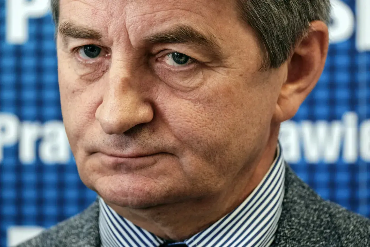 Mężczyzna w garniturze, w tle niebieska krata z białymi literami. Czy to on wyjaśnia, dlaczego posłowie PiS zostali oskarżeni o nadużywanie lotów rządowych?
