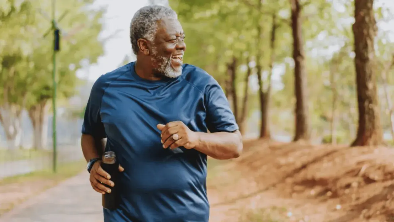 Un homme souriant court dans un parc, tenant une bouteille d'eau. Il est un exemple d'exercices pour perdre du ventre après 60 ans.