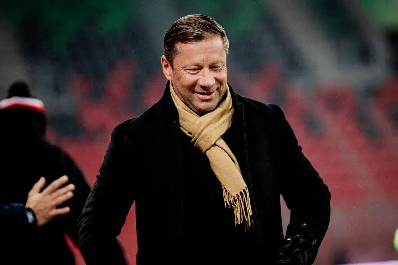 Uśmiechnięty Dariusz Banasik w czarnym płaszczu i beżowym szaliku, na tle trybun stadionu.