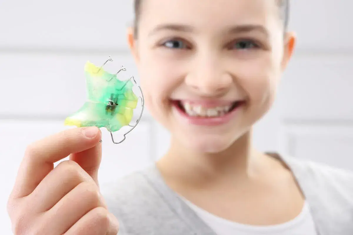 Niña sonriente muestra un retenedor dental verde y amarillo, listo para limpiar.