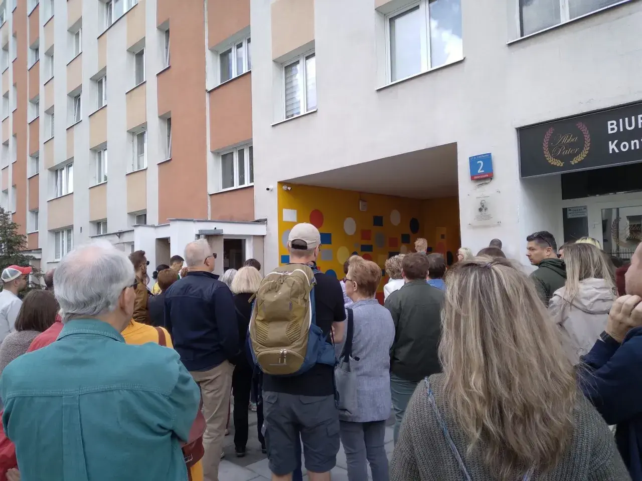 Grupa ludzi stoi przed blokiem mieszkalnym z kolorową ścianą i numerem 2.