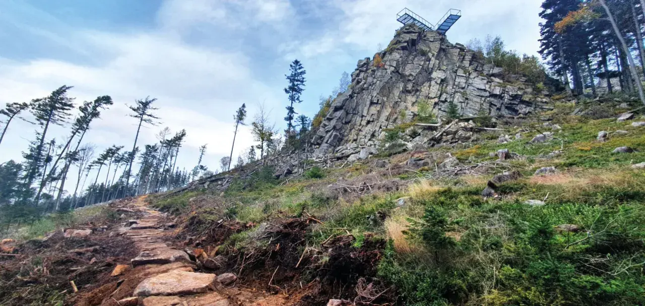 Kamienista ścieżka prowadzi ku skalistemu szczytowi z platformą widokową. W oddali widać las, a krajobraz przypomina ten z **Trójaka w Lądku Zdroju**.