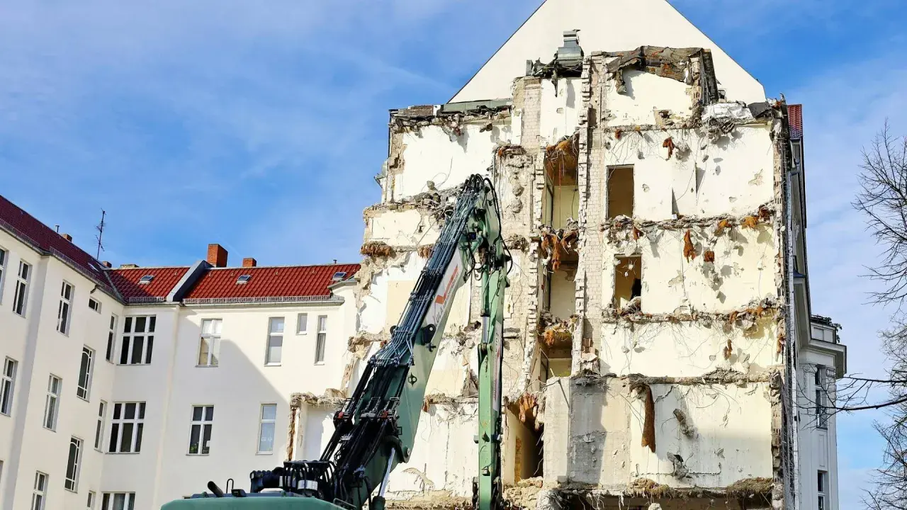 Koparka pracuje przy rozbiórce budynku, odsłaniając jego zniszczoną konstrukcję.