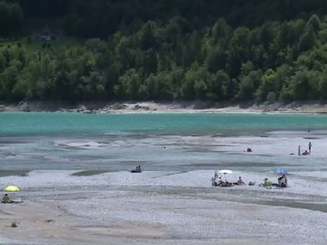Persone rilassate sulla spiaggia del lago di Barcis, con acque turchesi e boschi sullo sfondo.