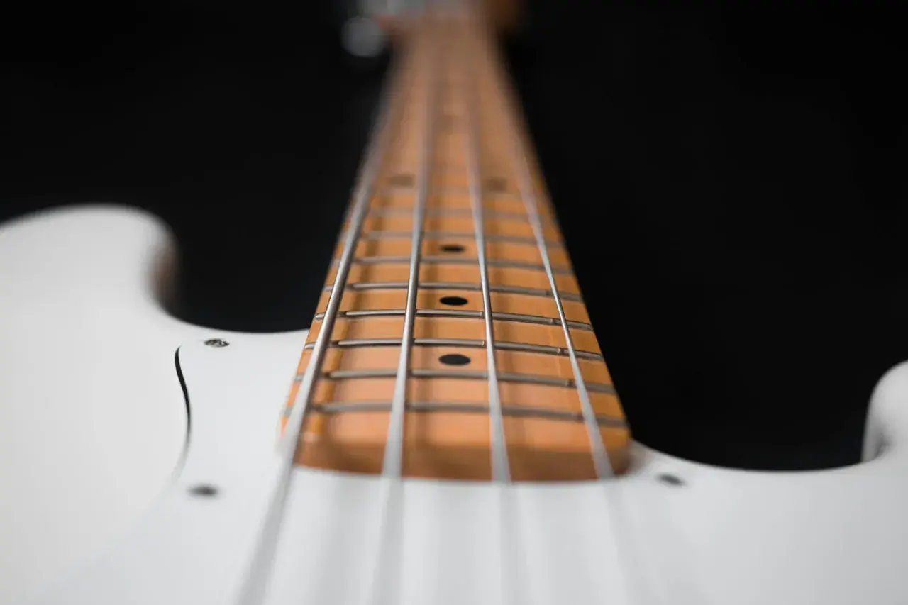 Primo piano del manico di un basso elettrico bianco, con le corde metalliche che corrono lungo la tastiera in legno chiaro.