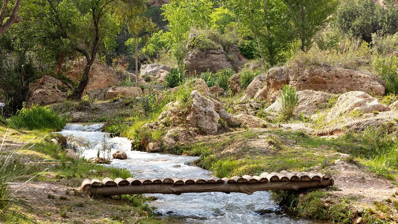 Un puente rústico cruza un arroyo en un paisaje rocoso y verde, evocando la serenidad de una cascada en Chelva.