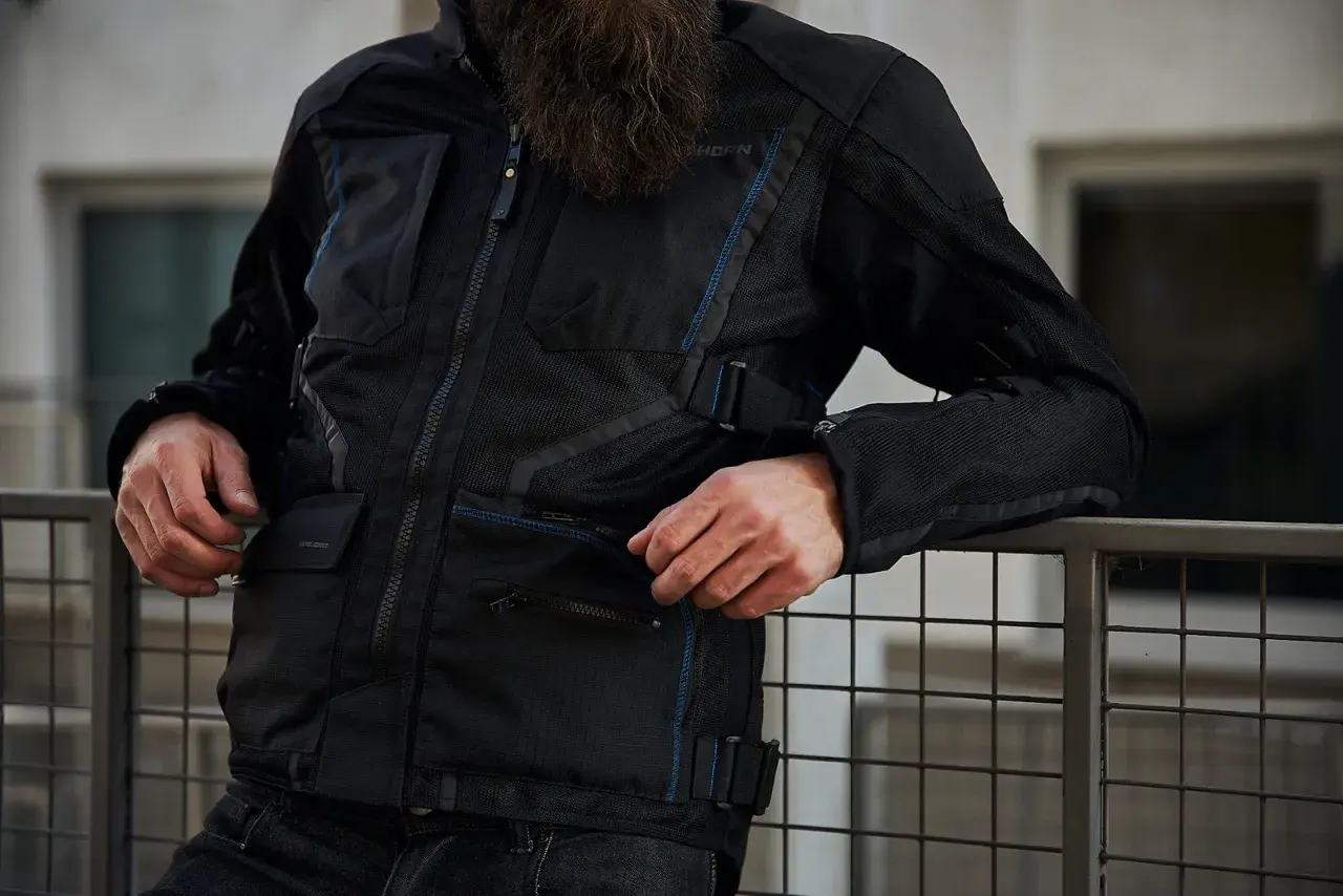 Mężczyzna w czarnej kurtce motocyklowej Rebelhorn Scandal II z niebieskimi akcentami, oparty o metalową barierkę.