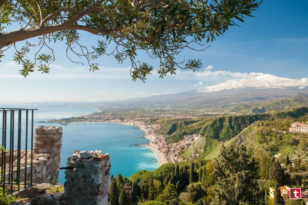 Vista panoramica di Naxos, Sicilia, con il mare azzurro, la costa e l'Etna innevato sullo sfondo, incorniciata da un ramo d'ulivo.