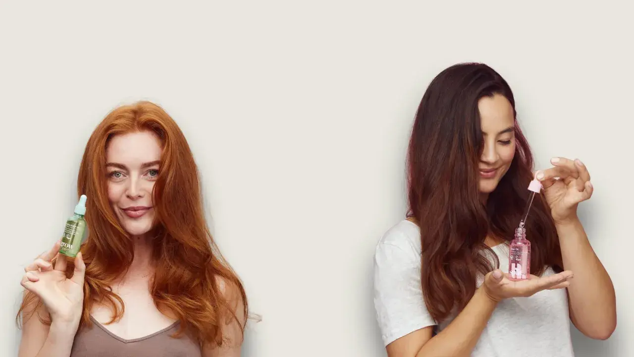 Dwie kobiety z buteleczkami olejków do włosów. Jedna z rudymi włosami trzyma zieloną buteleczkę, druga z brązowymi – różową. Idealne do maski z olejku rycynowego na mokre czy suche włosy.