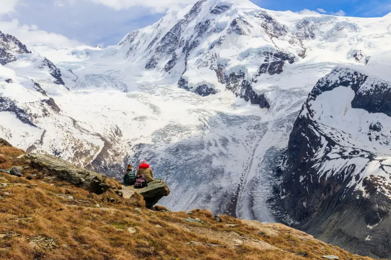 Two hikers rest on a rocky outcrop, gazing at the majestic, snow-covered peaks of the tour de Monte Rosa.