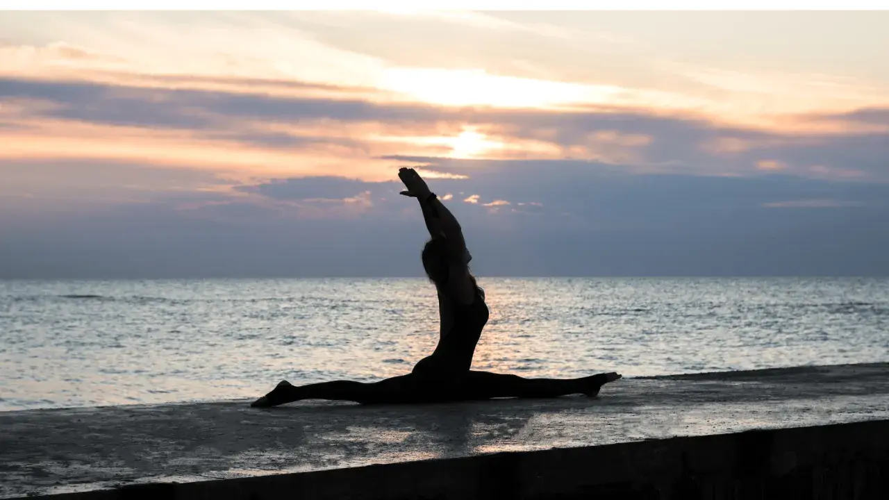 Silhouette di una donna in Ardha Hanumanasana sulla riva del mare al tramonto, con le braccia alzate verso il cielo.