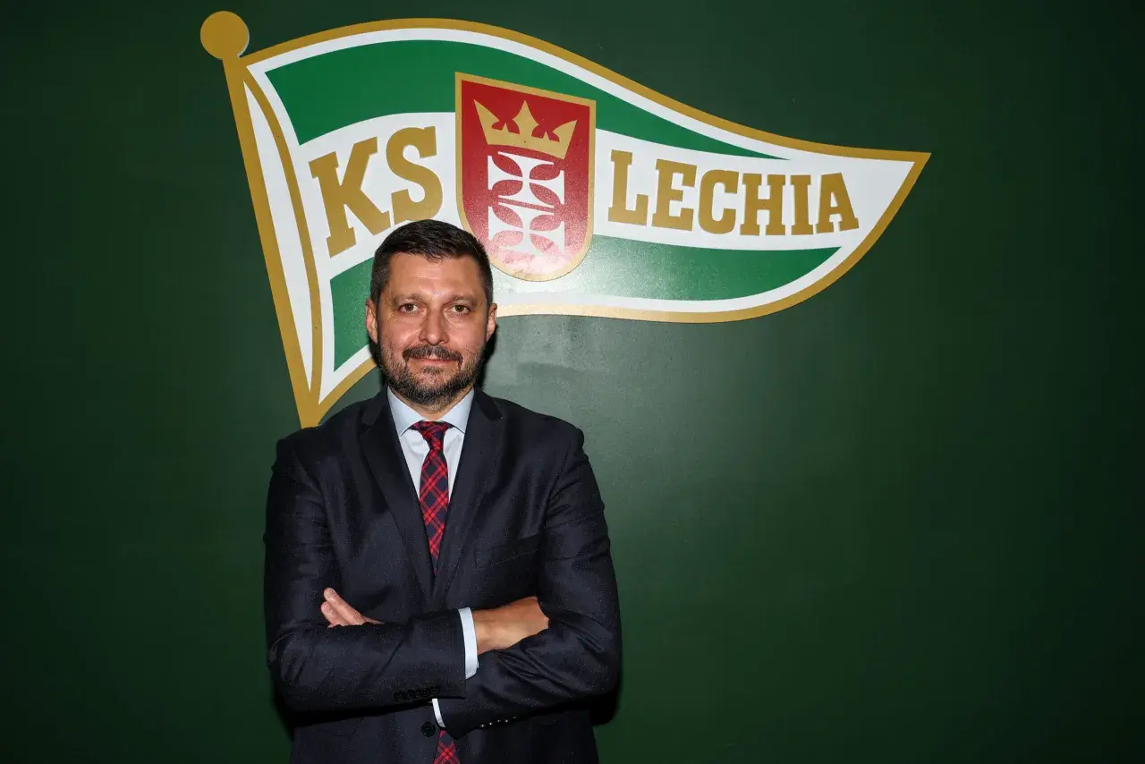 Marcin Kaczmarek, trener, stoi przed zieloną ścianą z logo Lechii Gdańsk.