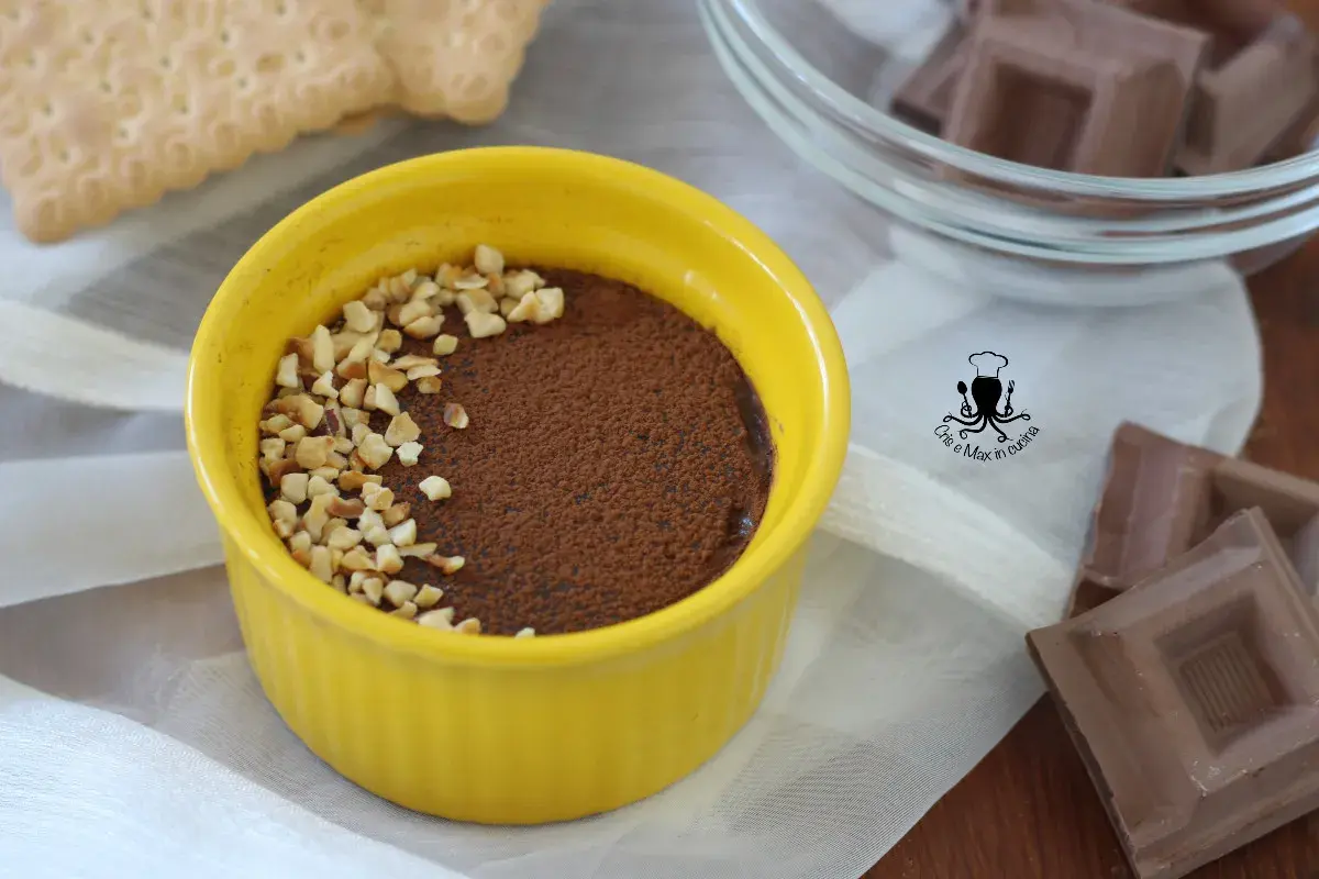 Budino al cioccolato in ciotola gialla, cosparso di cacao e nocciole tritate. Accanto, biscotti e cioccolato.