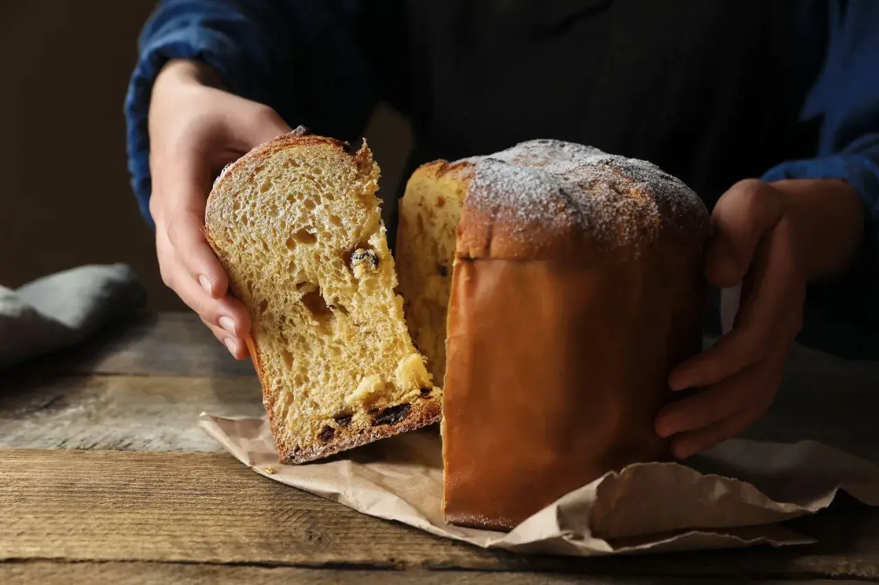 Puszysta babka drożdżowa włoska, z rodzynkami i kandyzowanymi owocami, posypana cukrem pudrem, prezentowana na drewnianym stole.