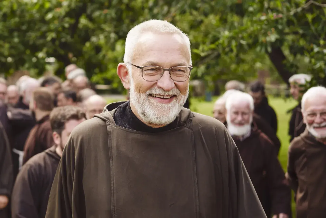 Bruder Paulus, ein älterer Mann mit Brille und Bart, lächelt in die Kamera. Hinter ihm stehen weitere Männer in braunen Kutten.