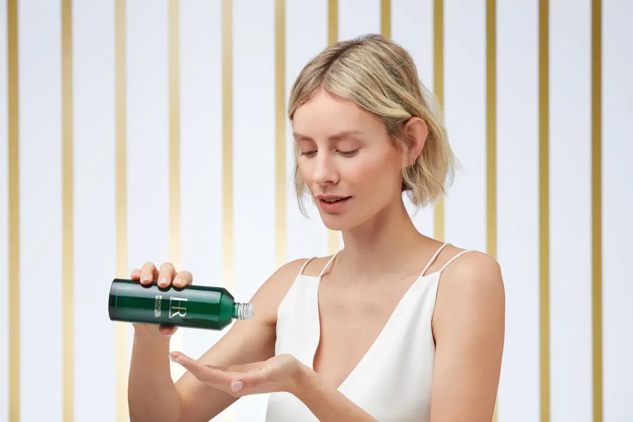 Frau mit blondem Bob gießt grünes Kosmetikprodukt aus Flasche in ihre Hand.