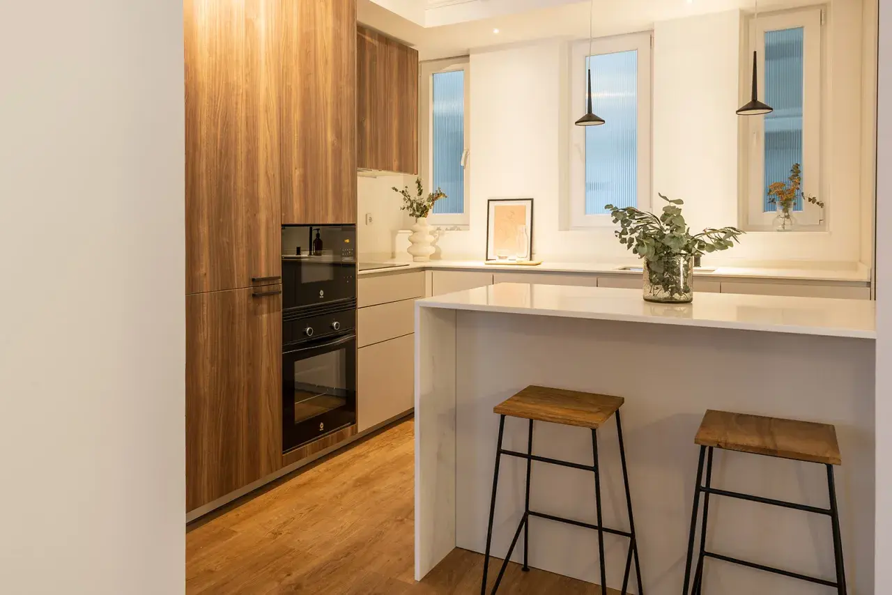 Cocina moderna con muebles de madera, encimera blanca y dos taburetes. La luz fría k ilumina el espacio, creando un ambiente acogedor.
