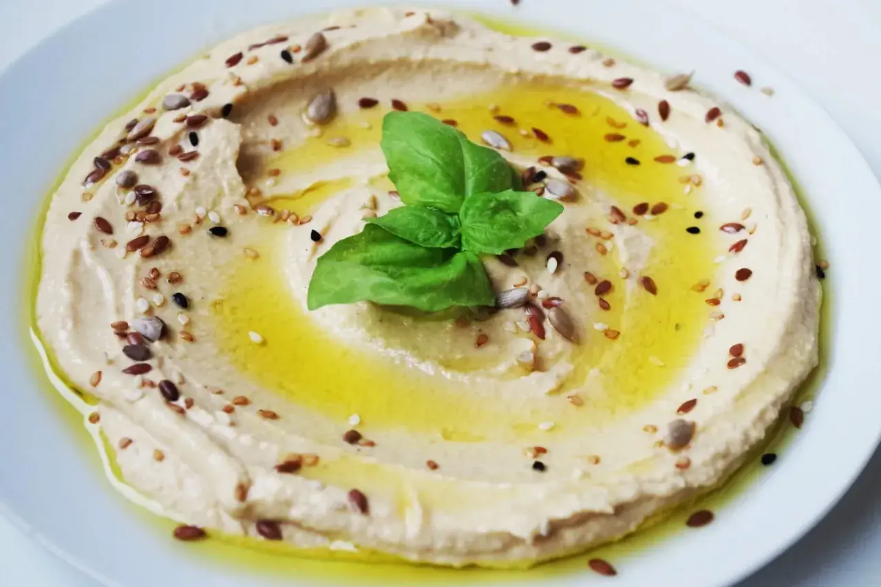 Hummus di ceci cremoso, condito con olio d'oliva, semi misti e una foglia di basilico. Perfetto da gustare con pane pita o verdure fresche.