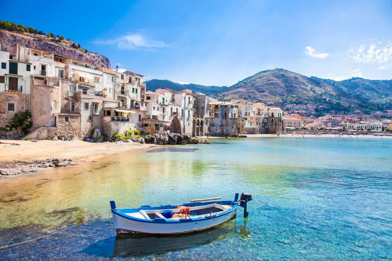 A charming fishing boat bobs in clear turquoise waters off the coast of Cefalù, a picturesque town with colorful buildings cascading down a hillside, a must-see among places to visit in Sicily.