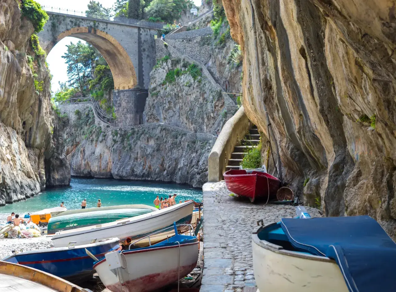 Boats line the shore of Fiordo di Furore, a stunning fjord with turquoise waters and a dramatic arched bridge overhead.
