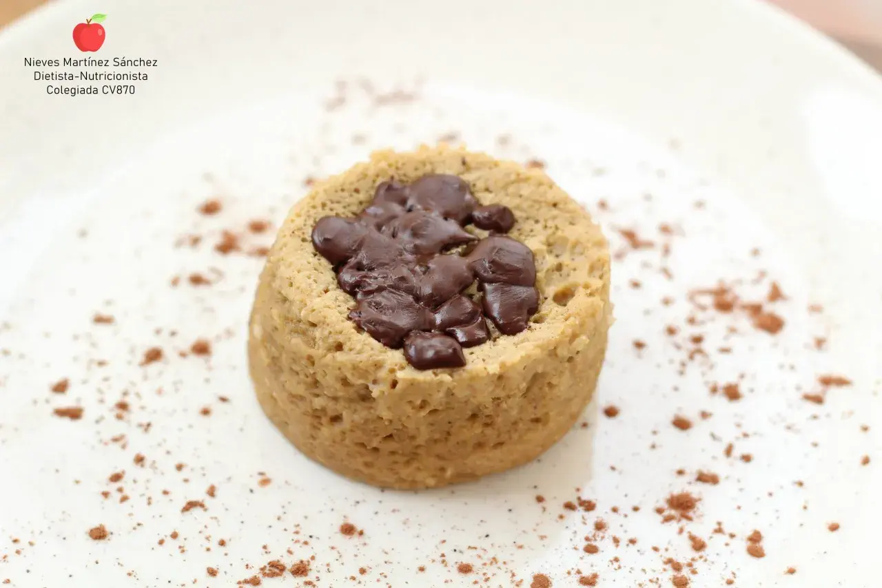Delicioso mug cake saludable con trozos de chocolate negro, espolvoreado con cacao en polvo sobre un plato blanco.