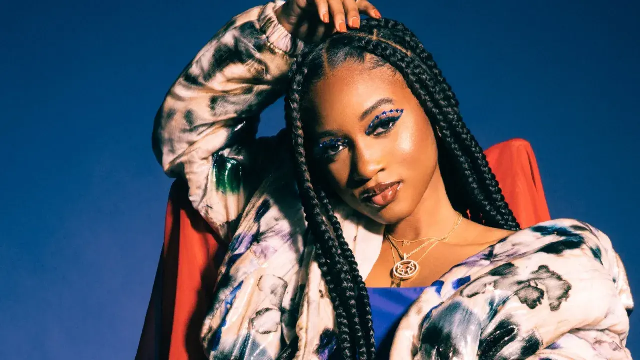 Nija Charles, with striking blue eye makeup and braids, poses confidently in a colorful jacket against a blue backdrop.