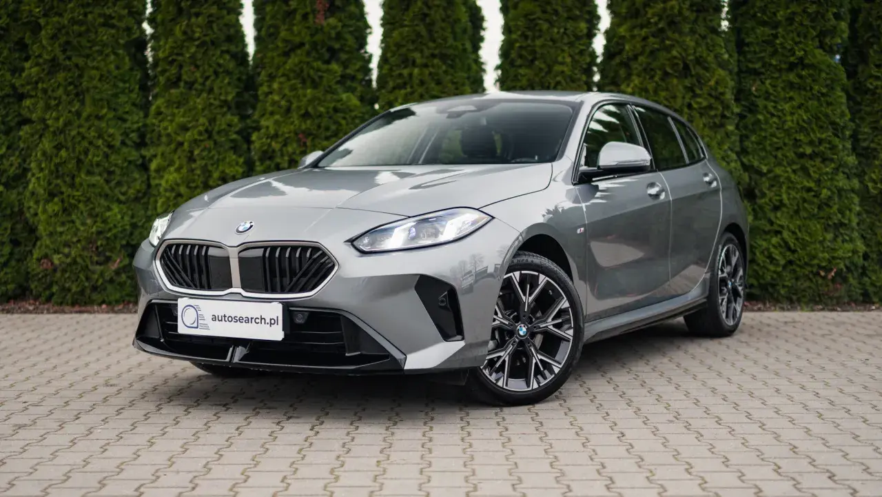 Szare BMW 118d z czarnym grillem i felgami stoi przed żywopłotem.