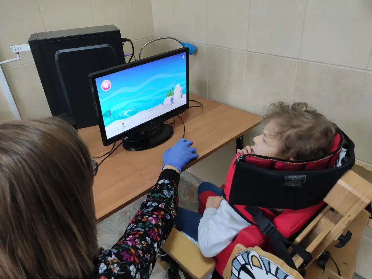Dziecko w specjalnym foteliku siedzi przed komputerem, na ekranie gra. Obok siedzi osoba w rękawiczce.