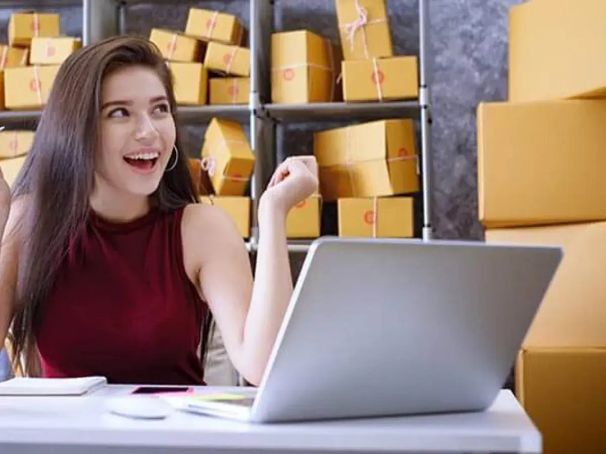 Szczęśliwa młoda kobieta przy laptopie, otoczona paczkami. To inspiracja, jak zacząć dropshipping i rozwijać swój biznes online.