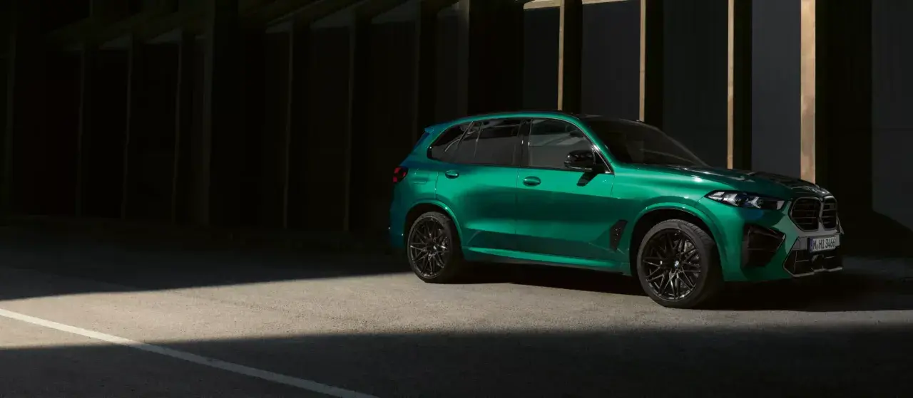 Zielony BMW X5 M z czarnymi felgami stoi na tle ciemnej architektury, oświetlony pasem światła.