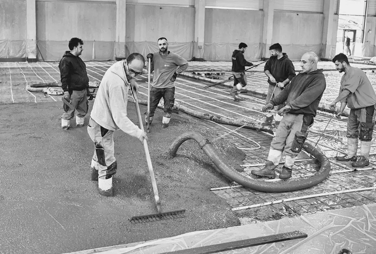 Arbeiter verteilen frisch gegossenen stahlfaserbeton bodenplatte mit Rechen. Beton wird aus Schlauch gepumpt.